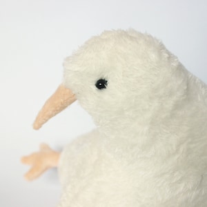 May include: A plush, off-white kiwi bird toy with a long, light brown beak and small black eyes. The bird has a soft, fluffy texture and a small, light brown foot is visible. The toy is set against a plain white background.