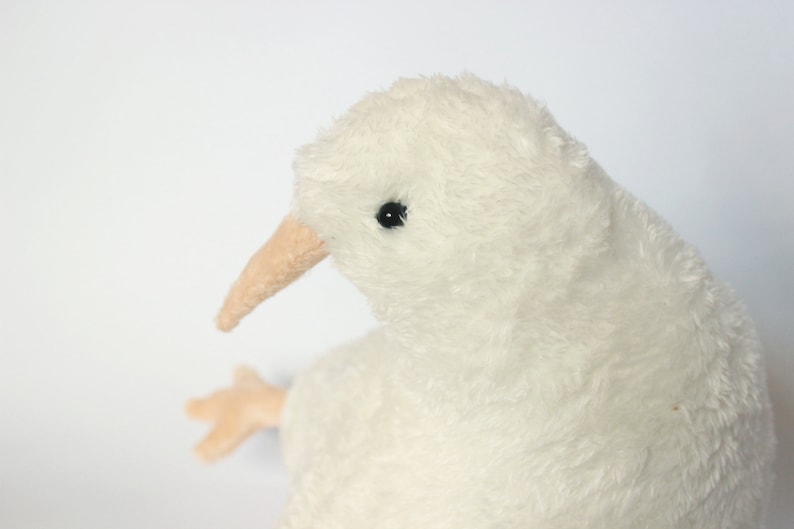May include: A plush, off-white kiwi bird toy with a long, light brown beak and small black eyes. The bird has a soft, fluffy texture and a small, light brown foot is visible. The toy is set against a plain white background.