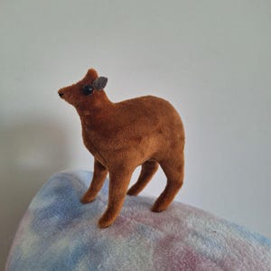 May include: A brown plush deer toy with small, dark grey ears and eyes. The deer stands on a soft, multi-coloured pillow with blue, pink, and white hues. The toy is made of a soft, fuzzy material.