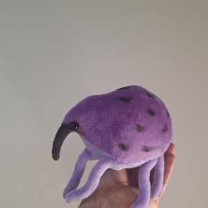 May include: A plush purple insect-like creature with black spots and a long, dark snout. The soft toy has four legs and is held in a hand. The toy is a whimsical design.