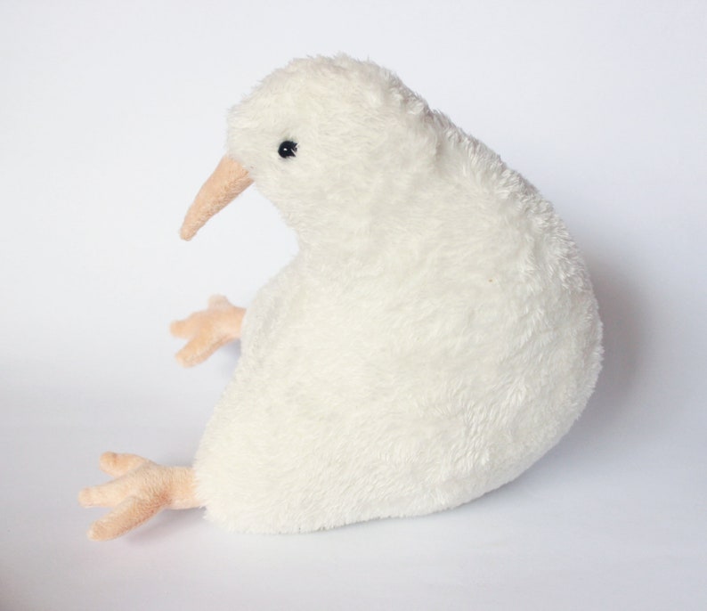 May include: A plush, off-white kiwi bird toy with a long, tan beak and small, tan feet. The soft, fluffy body is detailed with a small black eye. The toy is designed to sit upright, showcasing its unique shape and features.