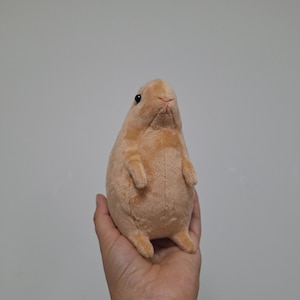 May include: A small, light brown plush toy shaped like a standing rodent. The stuffed animal features a smooth, soft texture, tiny front paws, short hind legs, and small black bead eyes with a stitched pink nose detail on its rounded face.