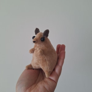 Op de afbeelding: Een klein, pluche, bruinachtig dier met donkerbruine oren en grote zwarte ogen. Het handgemaakte knuffeldier wordt in de hand van een persoon gehouden, wat de compacte grootte en zachte textuur laat zien. Het speelgoed heeft kleine armen en benen.