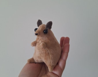 May include: A small, plush, tan-coloured animal with dark brown ears and large black eyes. The handmade stuffed animal is held in a person's hand, showcasing its compact size and soft texture. The toy has small arms and legs.