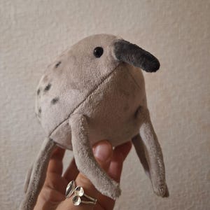 May include: A plush toy in the shape of a creature with a long beak and four legs. The toy is light gray with darker gray spots and a black beak. The toy has a stitched design and a black eye. The toy is being held by a person.