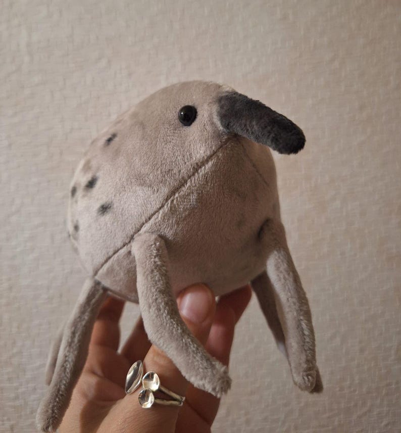 May include: A plush toy in the shape of a creature with a long beak and four legs. The toy is light gray with darker gray spots and a black beak. The toy has a stitched design and a black eye. The toy is being held by a person.