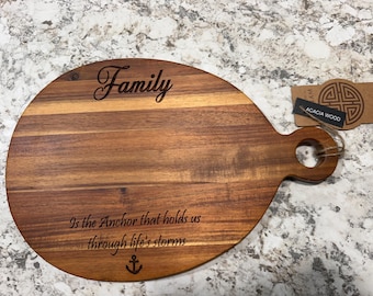 May include: An oval acacia wood serving board with a handle featuring a hole. The board is engraved with the text "Family" at the top and "Is the Anchor that holds us through life's storms" above a small anchor icon at the bottom.