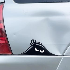 May include: A black vinyl car decal featuring a stylized character peeking out from behind a curved edge. The design shows a dark silhouette with a raised hand and two large, white, wide-eyed, suspicious-looking eyes peering forward.