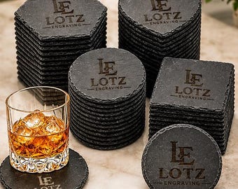 May include: A collection of black slate coasters in hexagonal, heart, square, and round shapes, each engraved with the text LE LOTZ ENGRAVING. A clear glass tumbler filled with amber liquid and ice cubes sits on one round coaster.