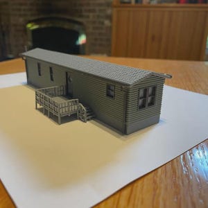 May include: A gray 3D printed miniature model of a mobile home featuring horizontal siding, a shingled roof, multiple rectangular windows, a single door, and an attached front deck with stairs and a railing, all rendered in a solid monochromatic finish.