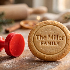 Könnte beinhalten: Ein runder, hellbrauner Keks mit dem geprägten Text The Miller Family in der Mitte, platziert neben einem leuchtend roten, runden Kunststoffstempel mit Griff, beide auf einer Holzoberfläche mit einer leichten Bestäubung aus Mehl.