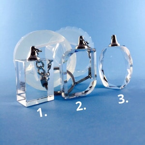 May include: Three clear glass keychains with metal rings and tops. The keychain on the left is rectangular, the middle keychain is square, and the keychain on the right is oval. The keychains are on a blue background.