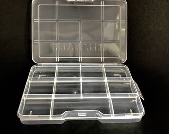May include: A clear plastic storage box with a hinged lid and 10 compartments. The lid has a ruler printed on the inside.