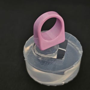 May include: A light purple ring with a rectangular band is displayed on a clear, circular mold. The ring has a smooth, glossy finish. The mold is made of a translucent material and sits on a dark surface.