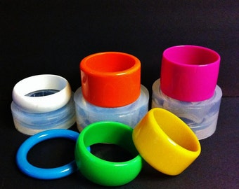 May include: Five colorful plastic bangles in white, orange, pink, yellow, and green. The bangles are arranged in a semi-circle on a white surface.