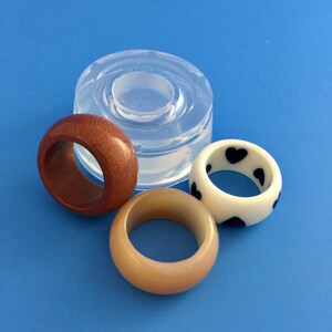 May include: Three resin rings, one brown, one beige, and one white with black hearts. The rings are sitting on a blue surface next to a clear silicone mold.