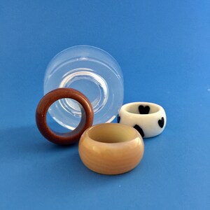 May include: Three resin rings, one brown, one beige, and one white with black hearts. The rings are sitting on a blue surface.