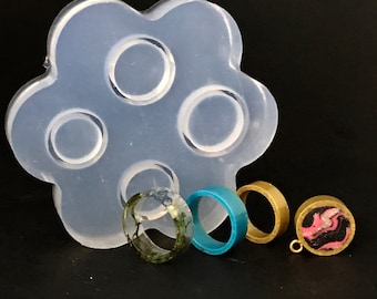 May include: A silicone mold with six circular cavities for making resin rings. Four rings are shown, two with clear resin and green accents, one with blue resin, and one with gold metal and pink and black resin.