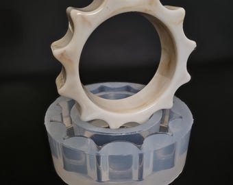 May include: A cream-coloured, sunburst-shaped bangle bracelet rests on a clear, circular mould. The bracelet has a glossy finish with subtle brown marbling. The mould is made of flexible, translucent silicone and has a matching sunburst pattern.