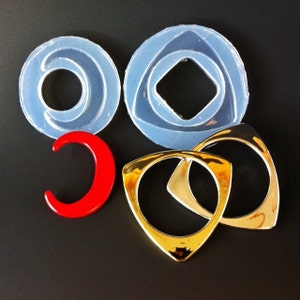 May include: Four silicone molds for making jewelry. The molds are in the shapes of a circle, a triangle, and two crescent moons. The molds are clear and have a shiny finish. There are also two gold-colored metal rings.