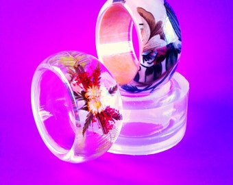 May include: Three resin rings with different designs. One ring is clear with pressed flowers, another is clear with a floral pattern, and the third is a wooden ring with a floral pattern.