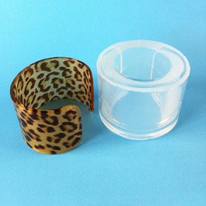 May include: A clear plastic mold for making resin bracelets. The mold is shaped like a wide bangle. Next to the mold is a finished resin bracelet with a leopard print design.