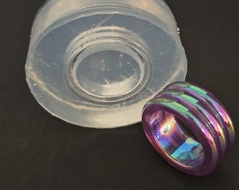 May include: A clear, cylindrical mold next to a purple and iridescent ring. The ring has a rectangular shape with horizontal ridges. The mold appears to be made of silicone or a similar material.