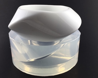 May include: A white ceramic bowl with a unique, twisted design sits inside a clear silicone mould. The mould is also twisted, creating a matching shape for the bowl.