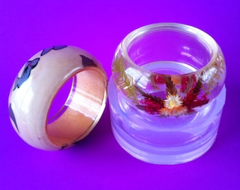 May include: Two clear resin bracelets with a floral design. One bracelet is white with a black and white design, the other is clear with dried flowers.