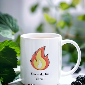 Puede incluir: Una taza de cerámica blanca con un gráfico de una llama estilizada en rojo, naranja y amarillo. Debajo de la llama, el texto dice: "You make life warm!". La taza tiene un asa estándar y un cuerpo cilíndrico.