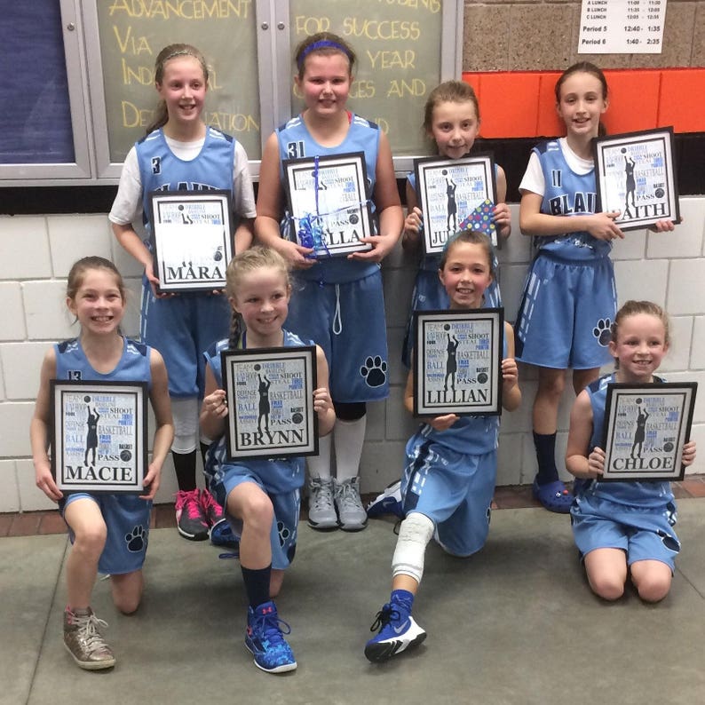 May include: A group of young girls in blue basketball uniforms pose with framed certificates. Each certificate features a basketball player silhouette and the words "Team, Dribble, Shoot, Basketball, Pass" with a girl's name at the bottom. The certificates are framed in black.