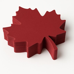 May include: A stack of burgundy-colored paper napkins, each cut in the shape of a maple leaf. The napkins are neatly arranged, showcasing the detailed leaf outline. The image is set against a plain white background.