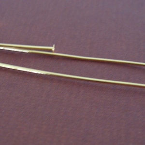 May include: Close-up of several gold-colored head pins. The pins have a small, flat head and a long, slender shaft. They are scattered across a brown surface, with some pins in focus and others blurred in the background. These pins are used for jewelry making.