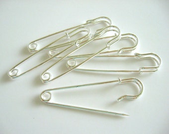 May include: A collection of five silver-toned metal safety pins arranged in a loose pile. Each pin features a pointed end, a coiled spring mechanism at the base, and a protective clasp that secures the sharp tip when closed.