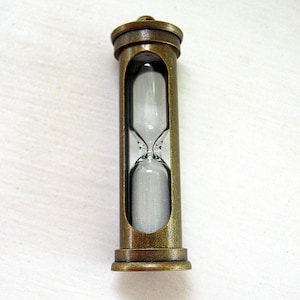 May include: An antique-style hourglass with a bronze-colored metal frame. The glass chamber holds white sand, visible through the open sides of the frame. The hourglass is designed with a classic, vintage aesthetic.