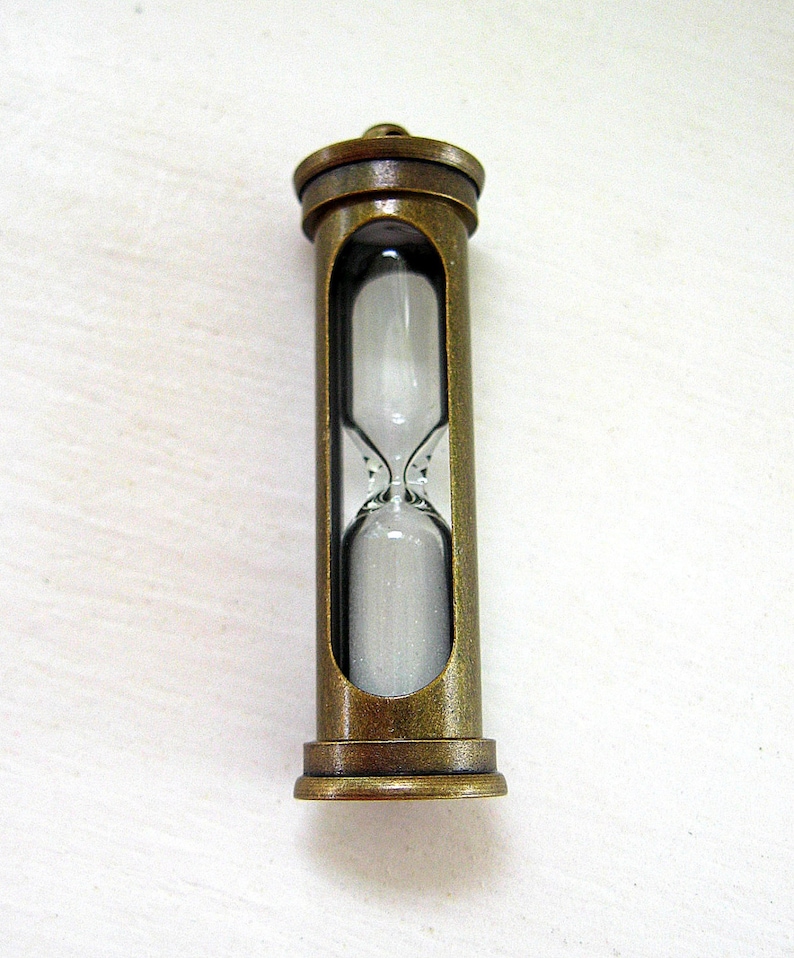 May include: An antique-style hourglass with a bronze-colored metal frame. The glass chamber holds white sand, visible through the open sides of the frame. The hourglass is designed with a classic, vintage aesthetic.