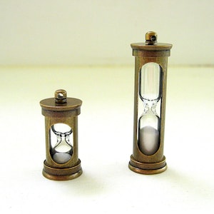 May include: Two antique-style hourglasses with bronze-colored metal frames. The glass chambers hold white sand. One hourglass is taller than the other. The hourglasses are decorative items, possibly for a desk or shelf.