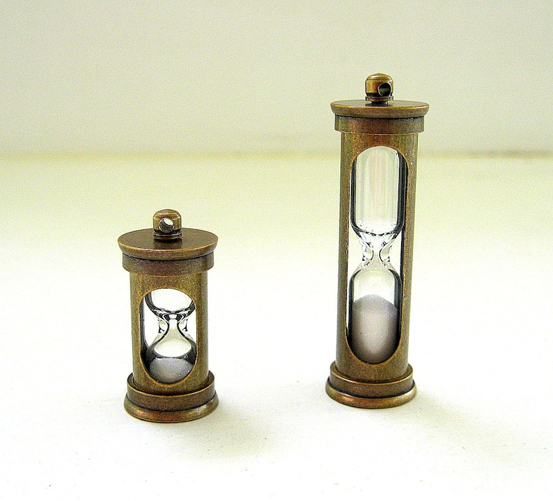 May include: Two antique-style hourglasses with bronze-colored metal frames. The glass chambers hold white sand. One hourglass is taller than the other. The hourglasses are decorative items, possibly for a desk or shelf.