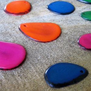 May include: A collection of colorful, teardrop-shaped pendants in various shades including pink, orange, blue, and green. Each pendant has a small hole at the top and a dark border. The pendants are arranged on a textured, neutral-toned surface.