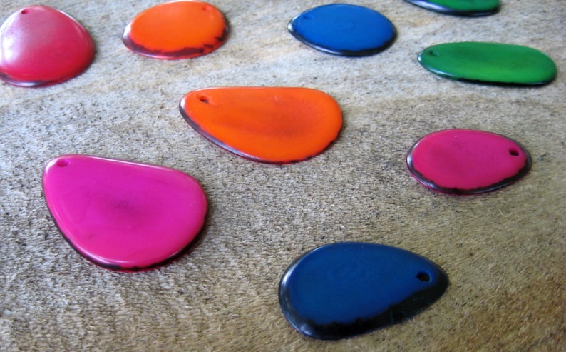 May include: A collection of colorful, teardrop-shaped pendants in various shades including pink, orange, blue, and green. Each pendant has a small hole at the top and a dark border. The pendants are arranged on a textured, neutral-toned surface.