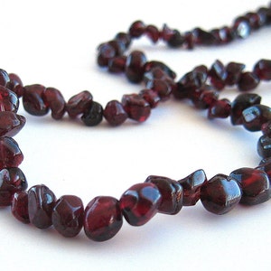 May include: A necklace made of dark red, irregularly shaped garnet beads. The beads are strung together, forming a circular shape. The background is plain white, highlighting the deep color of the gemstones.
