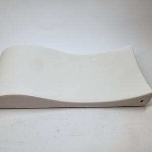 Puede incluir: Una bandeja de cerámica blanca y curvada con una superficie lisa. La bandeja tiene un diseño ondulado a lo largo de un borde y un pequeño agujero en la esquina. La forma general es rectangular, con esquinas redondeadas.