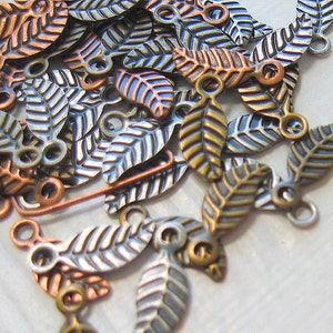 May include: A collection of small metal leaf charms in various colors, including copper, bronze, and silver. Each leaf has a detailed vein pattern and a small loop for attaching to jewelry or other crafts.