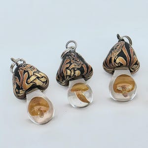May include: Three mushroom-shaped pendants featuring black and tan marbled caps with intricate swirling patterns. Each pendant has a clear, transparent stem containing a small, suspended amber-colored mushroom detail and a silver-toned metal jump ring.