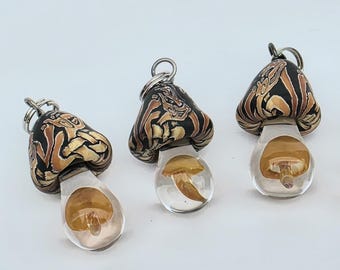 May include: Three mushroom-shaped pendants featuring black and tan marbled caps with intricate swirling patterns. Each pendant has a clear, transparent stem containing a small, suspended amber-colored mushroom detail and a silver-toned metal jump ring.