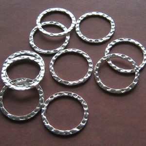 May include: Silver-toned metal hammered rings. The rings have a textured surface and a circular shape. The rings are scattered on a brown surface. These rings are suitable for jewelry making and craft projects.