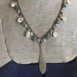 Puede incluir: Un collar con un colgante de pescado plateado, cuentas azules y blancas. El collar está en un maniquí.