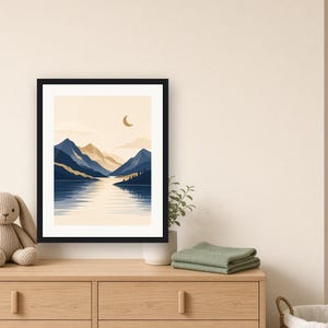 May include: A framed wall art print featuring a landscape illustration of dark blue mountains, a calm lake, and a crescent moon against a light beige sky. The artwork is enclosed in a simple black frame, showcasing a serene nature scene with layered peaks and reflections on the water surface.