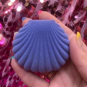 May include: A blue seashell-shaped jewelry box with a textured surface. The box is held against a background of pink and gold sequins. The box is designed to hold small jewelry items.