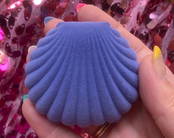 May include: A blue seashell-shaped jewelry box with a textured surface. The box is held against a background of pink and gold sequins. The box is designed to hold small jewelry items.